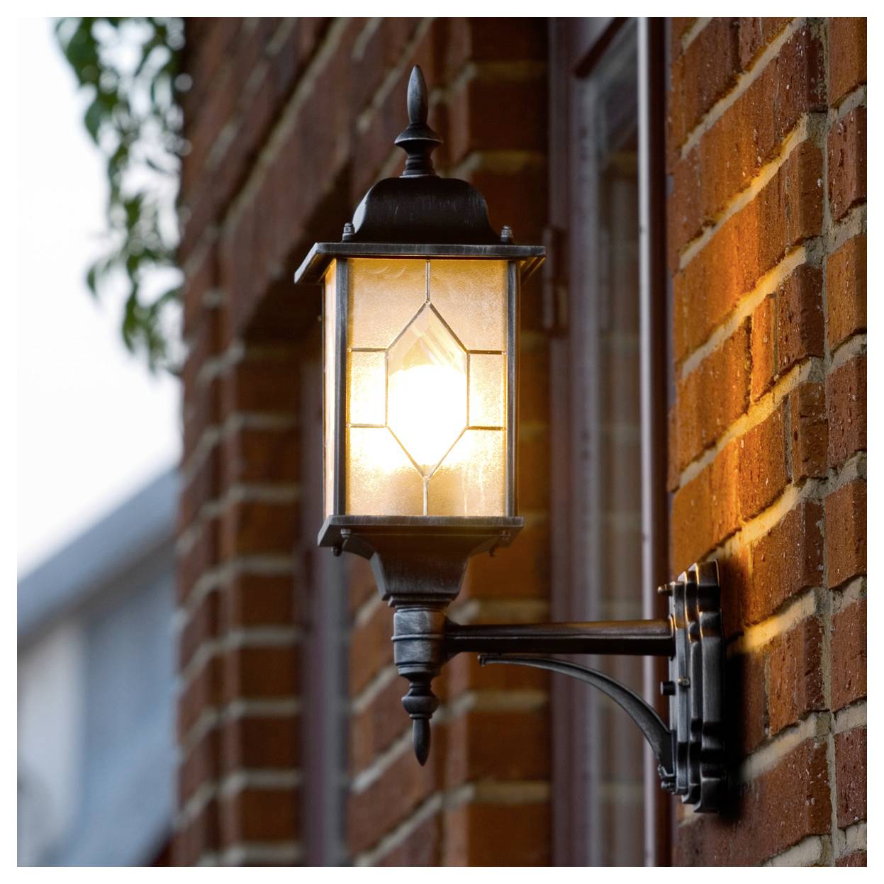 Lanterne en métal noir brillant montée sur un mur de briques à côté d'une fenêtre, projetant une lumière chaleureuse à l'extérieur.