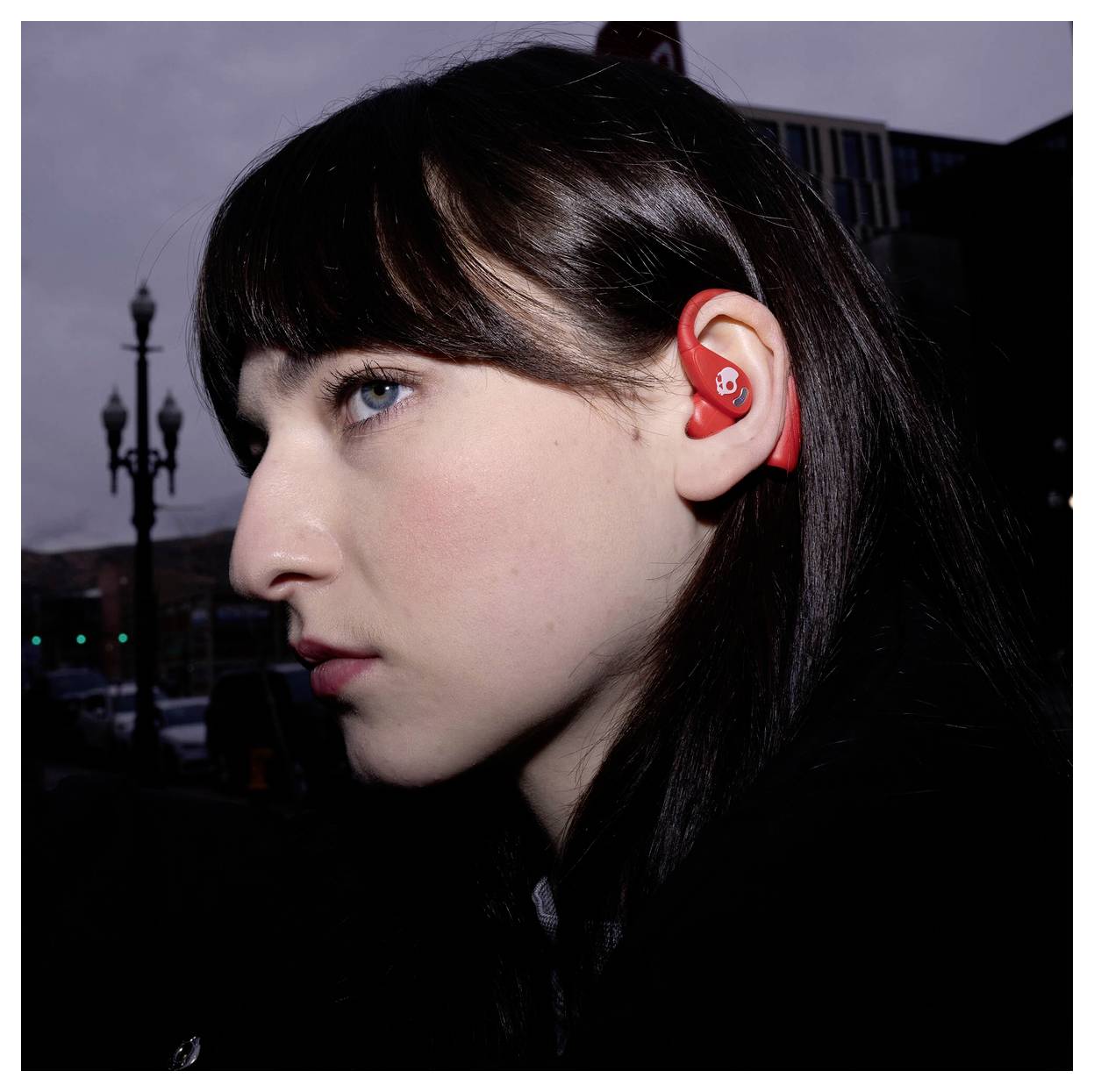 Gros plan de profil latéral d'une jeune femme aux cheveux noirs portant un écouteur rouge, en extérieur au crépuscule avec un réverbère flou et un bâtiment derrière elle.
