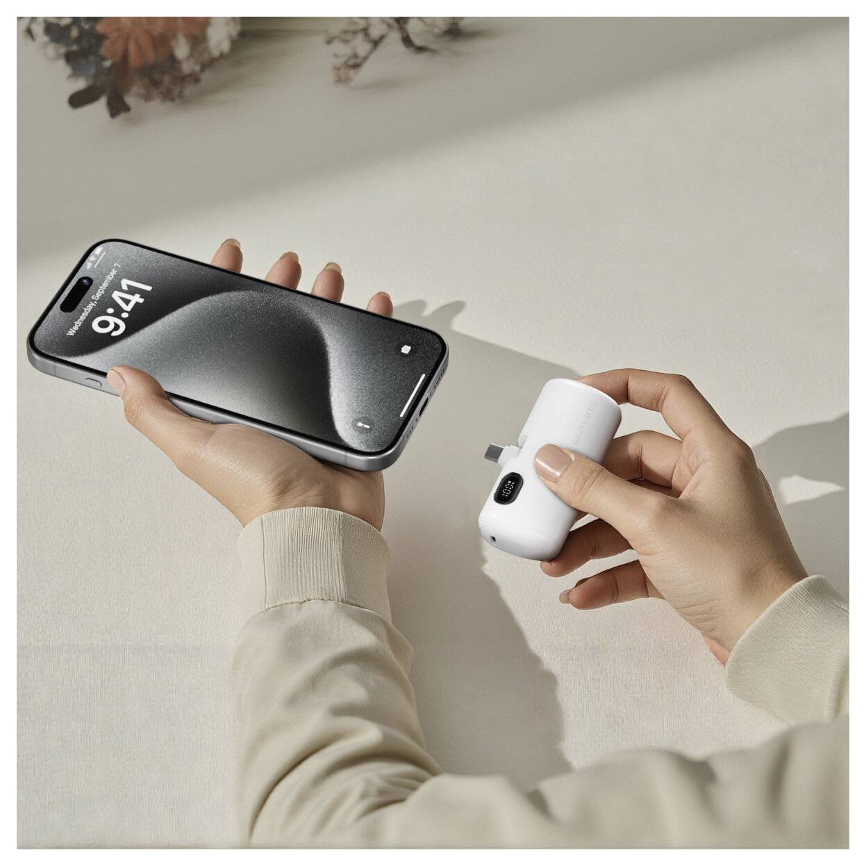 Mains tenant un smartphone et un petit adaptateur de charge sans fil blanc, suggérant une charge mobile sur une surface claire.