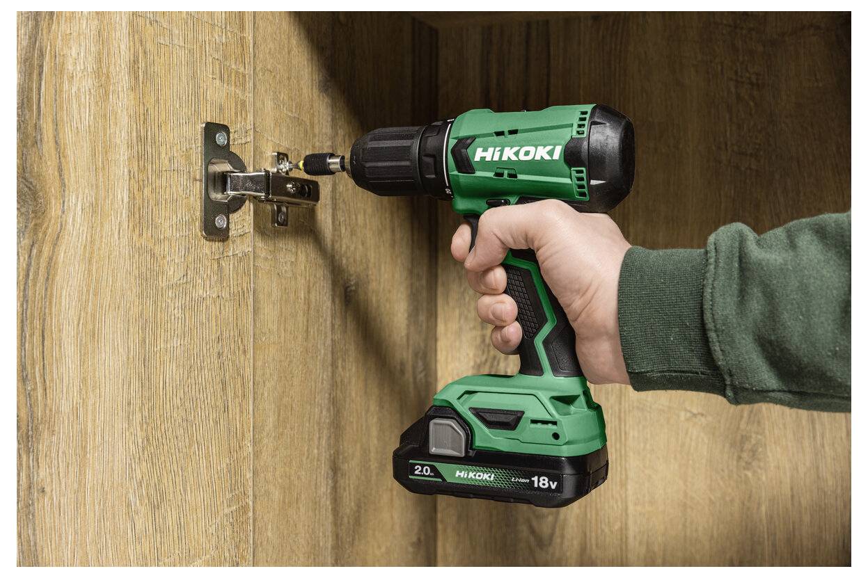 Une main utilisant une perceuse sans fil verte Hikoki pour serrer une charnière de placard sur un panneau de porte en bois.