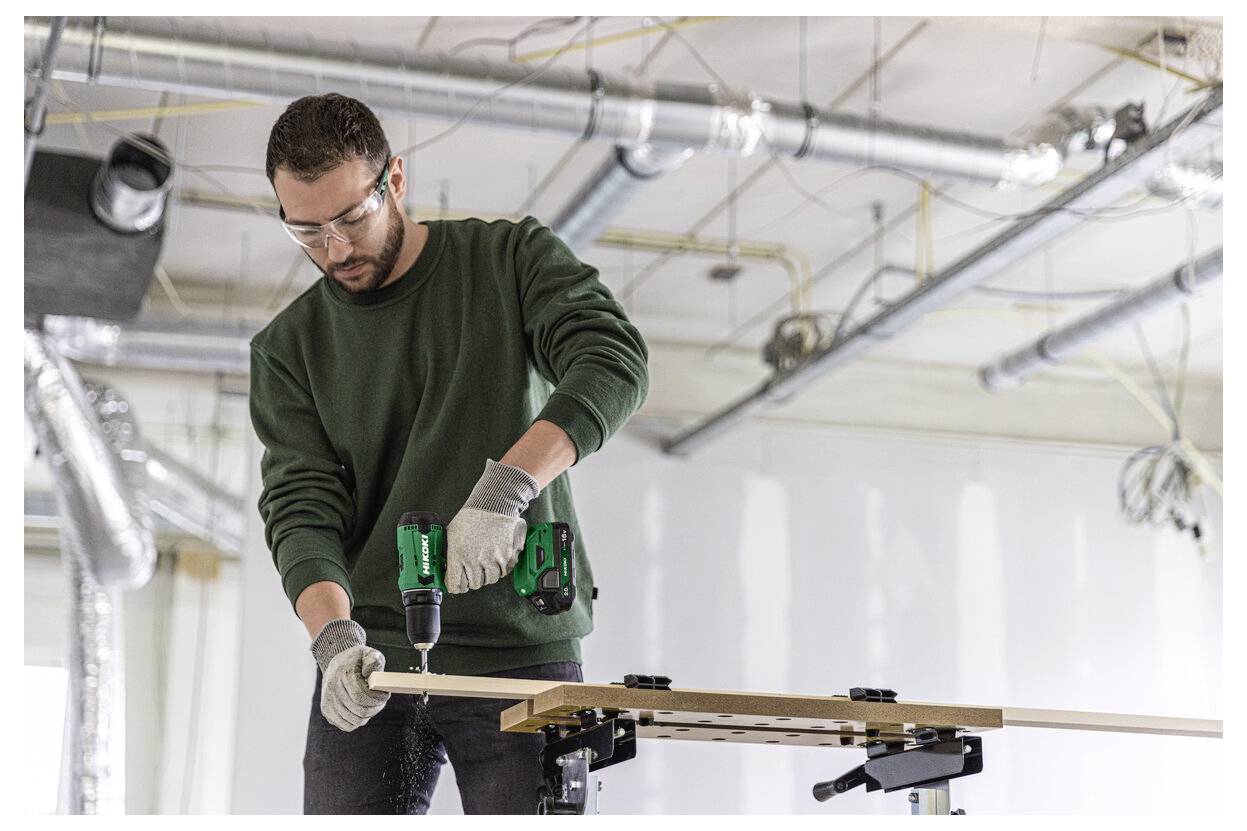 Un homme portant des lunettes de sécurité et des gants utilise une perceuse sans fil sur une planche en bois dans un atelier.
