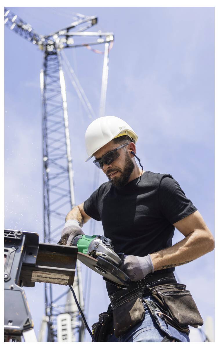 Ouvrier de la construction portant un casque de sécurité et des lunettes de protection utilisant une scie électrique sur un chantier de construction, avec une grue en arrière-plan.