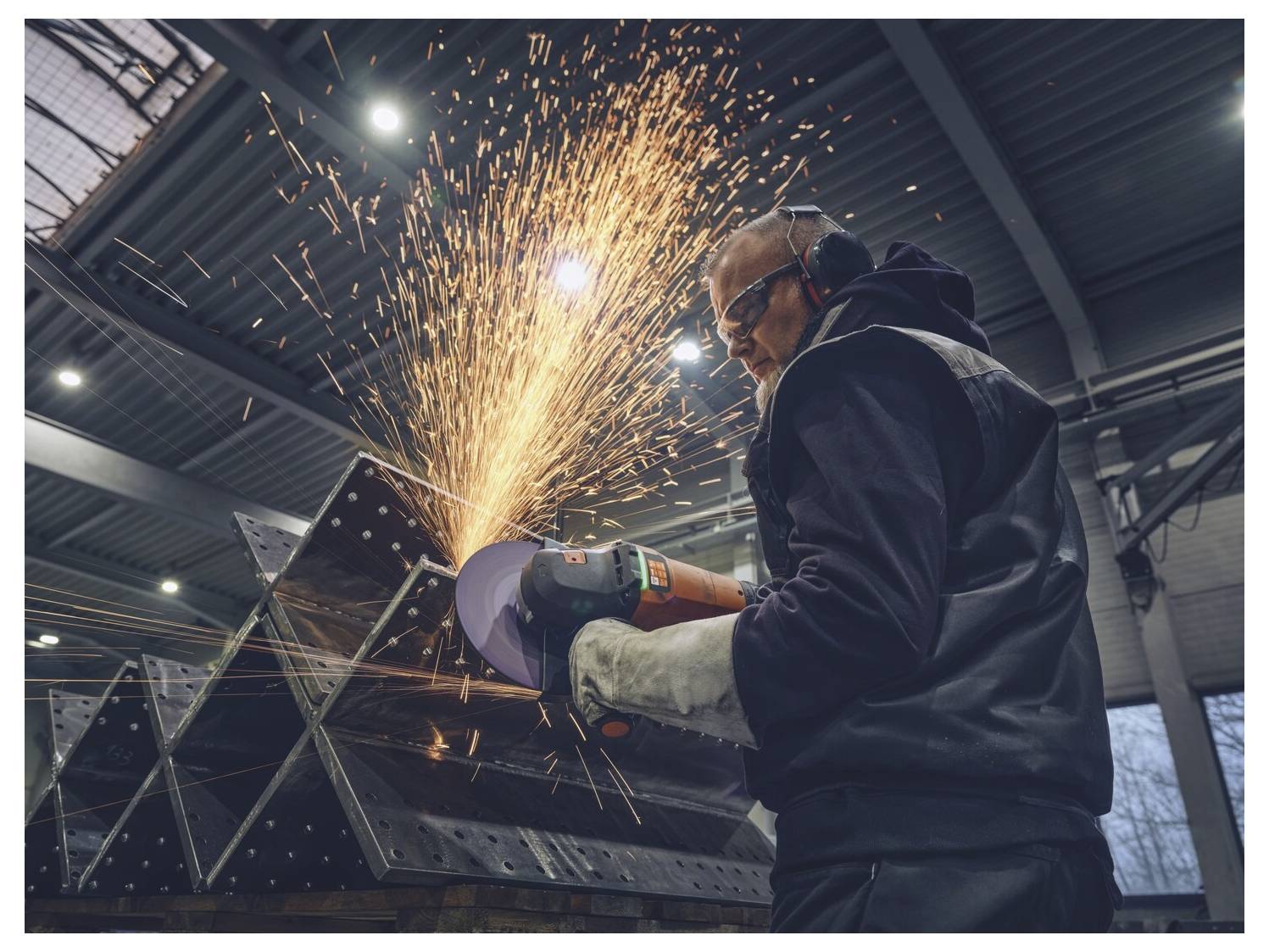 Un ouvrier utilisant une meuleuse d'angle sur du métal dans un atelier, produisant des étincelles brillantes tout en portant des lunettes de sécurité, une protection auditive et des gants.