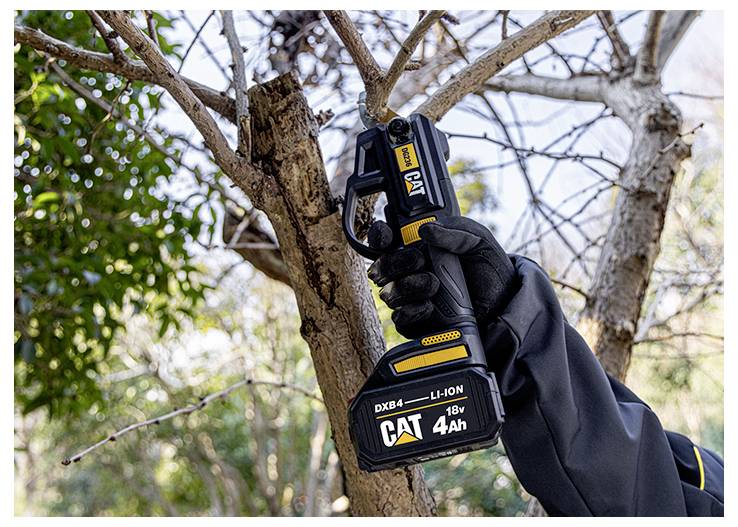 Main gantée utilisant une scie à élaguer sans fil CAT pour couper une branche d'arbre à l'extérieur parmi des branches dénudées.