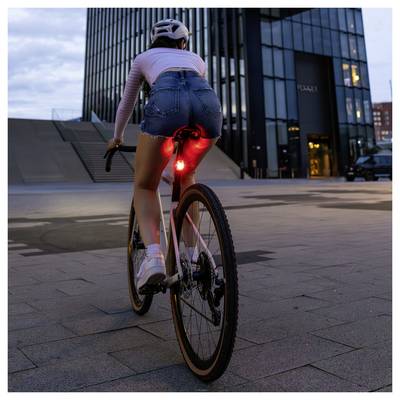 Une personne fait du vélo au crépuscule en ville ; un feu arrière rouge brille, des immeubles de bureaux modernes en arrière-plan.