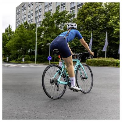 Un cycliste vêtu de bleu pédalant sur un vélo de route turquoise dans une rue urbaine, vu de dos près des arbres et des immeubles d'habitation.