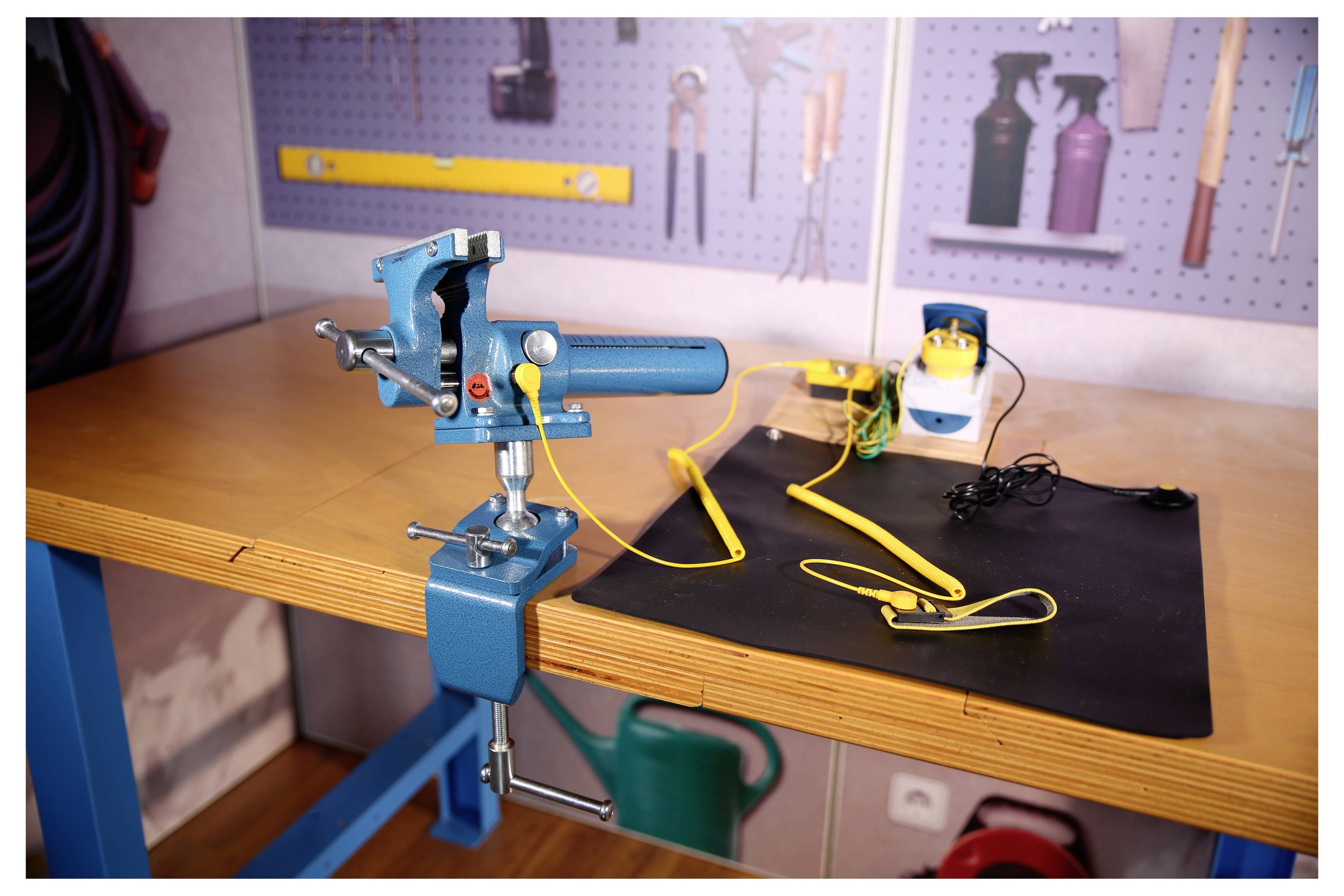 Étau d'établi bleu maintenant un outil cylindrique bleu sur un établi, avec des cordons jaunes et un petit appareil à côté dans un atelier.