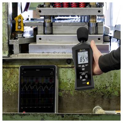 Une personne mesure le bruit d'une machine industrielle avec un sonomètre portatif tandis qu'une tablette affiche un graphique du niveau sonore.