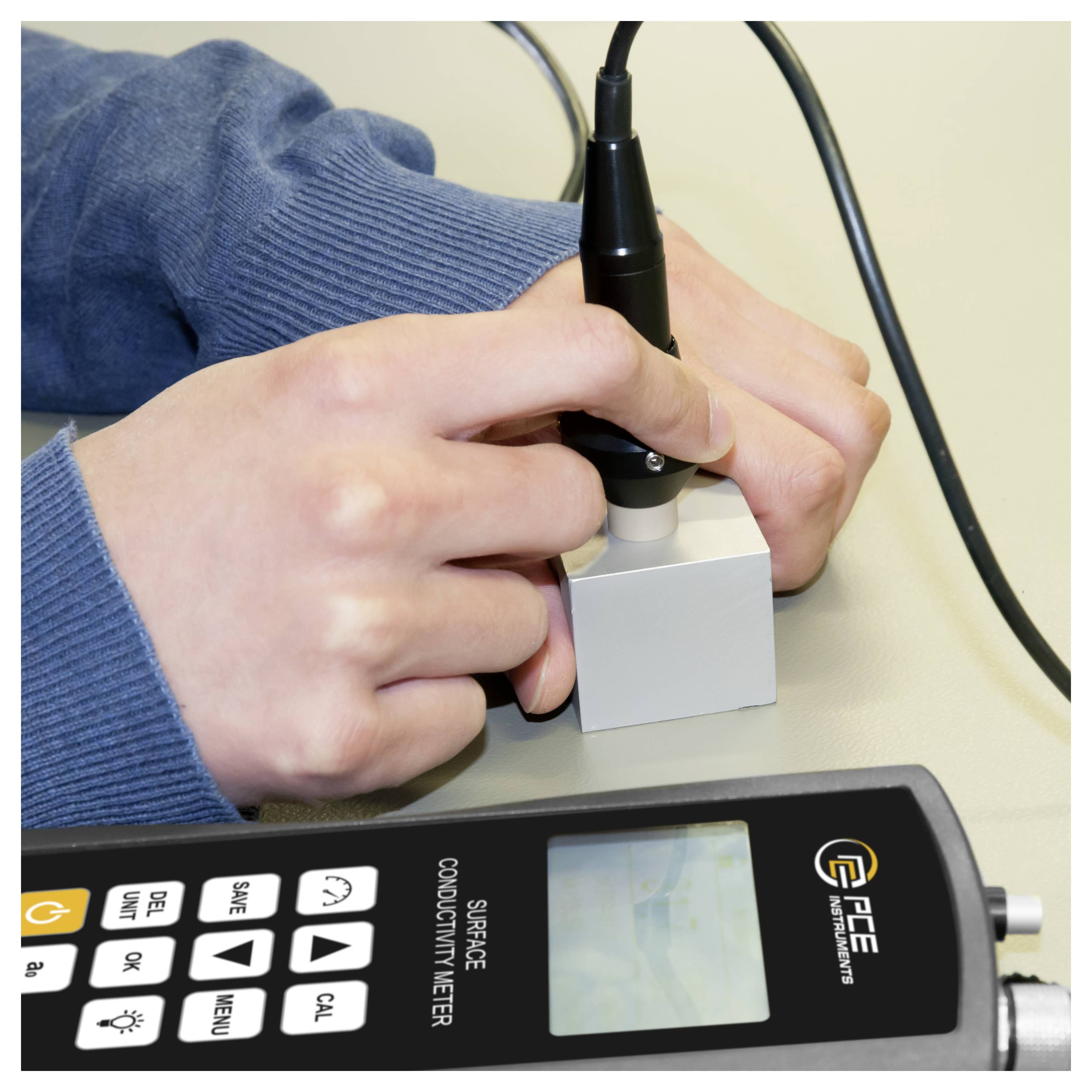 Des mains utilisant une sonde sur un appareil de mesure de conductivité de surface avec un écran numérique et des boutons de commande.