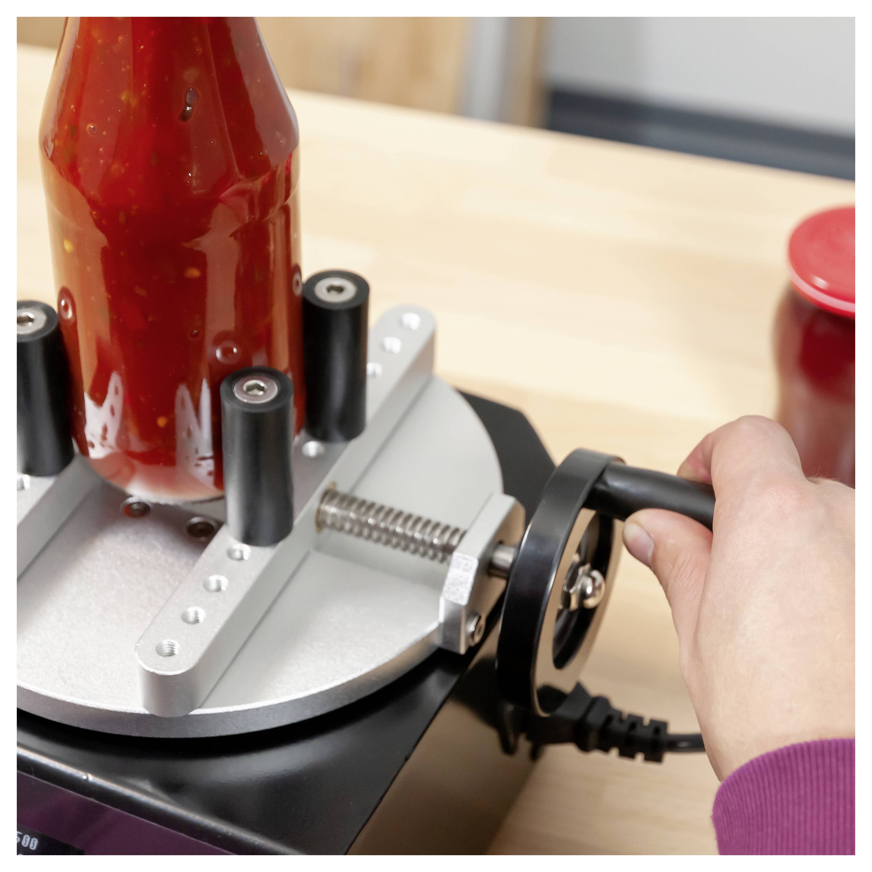 Une main tournant une roue de serrage pour fixer une bouteille rouge dans une machine de capsulage ou d'étiquetage de bouteilles.
