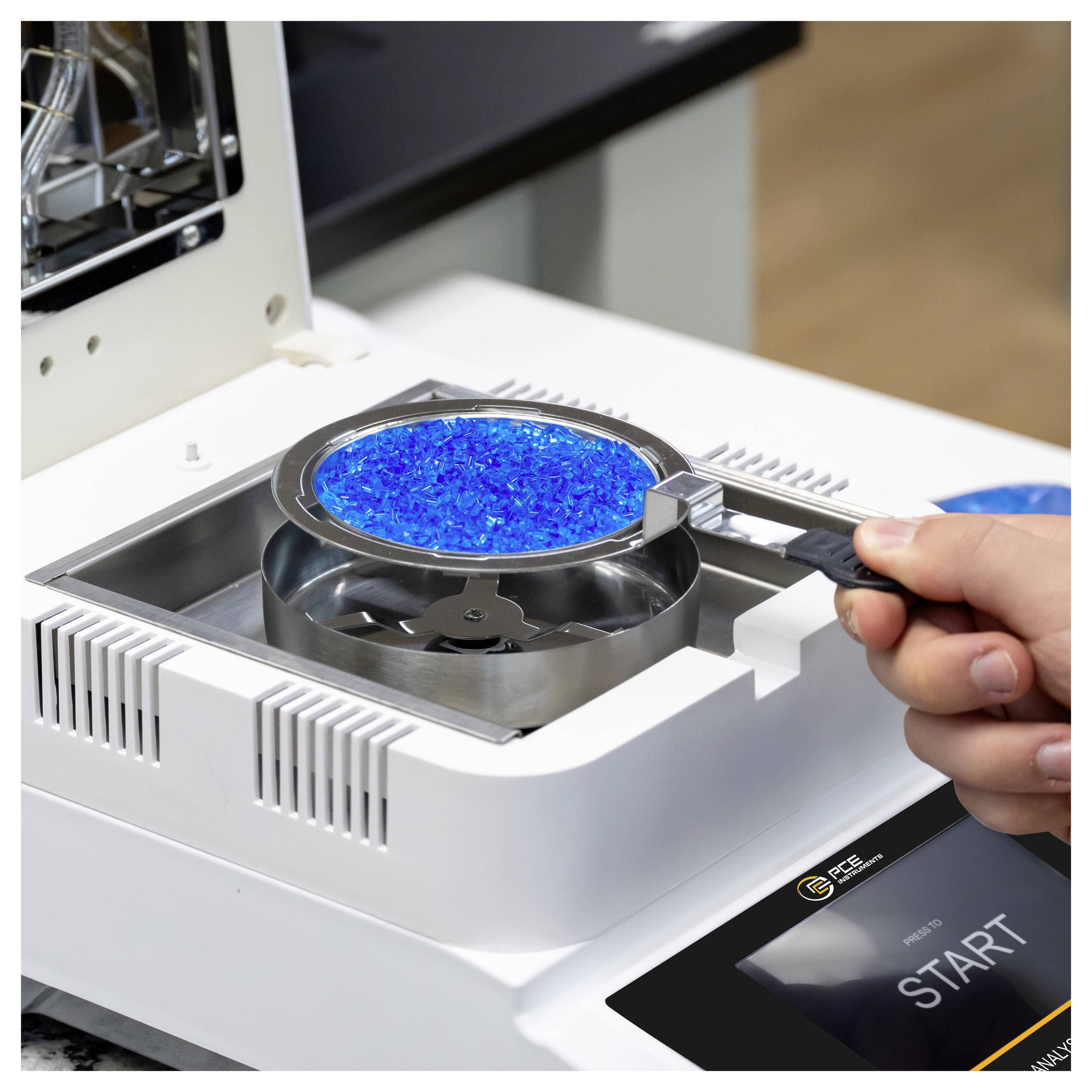 Une main tient une casserole de granules bleus au-dessus d'un analyseur de laboratoire avec un bouton DÉMARRER, ce qui suggère la préparation d'échantillons ou les tests.