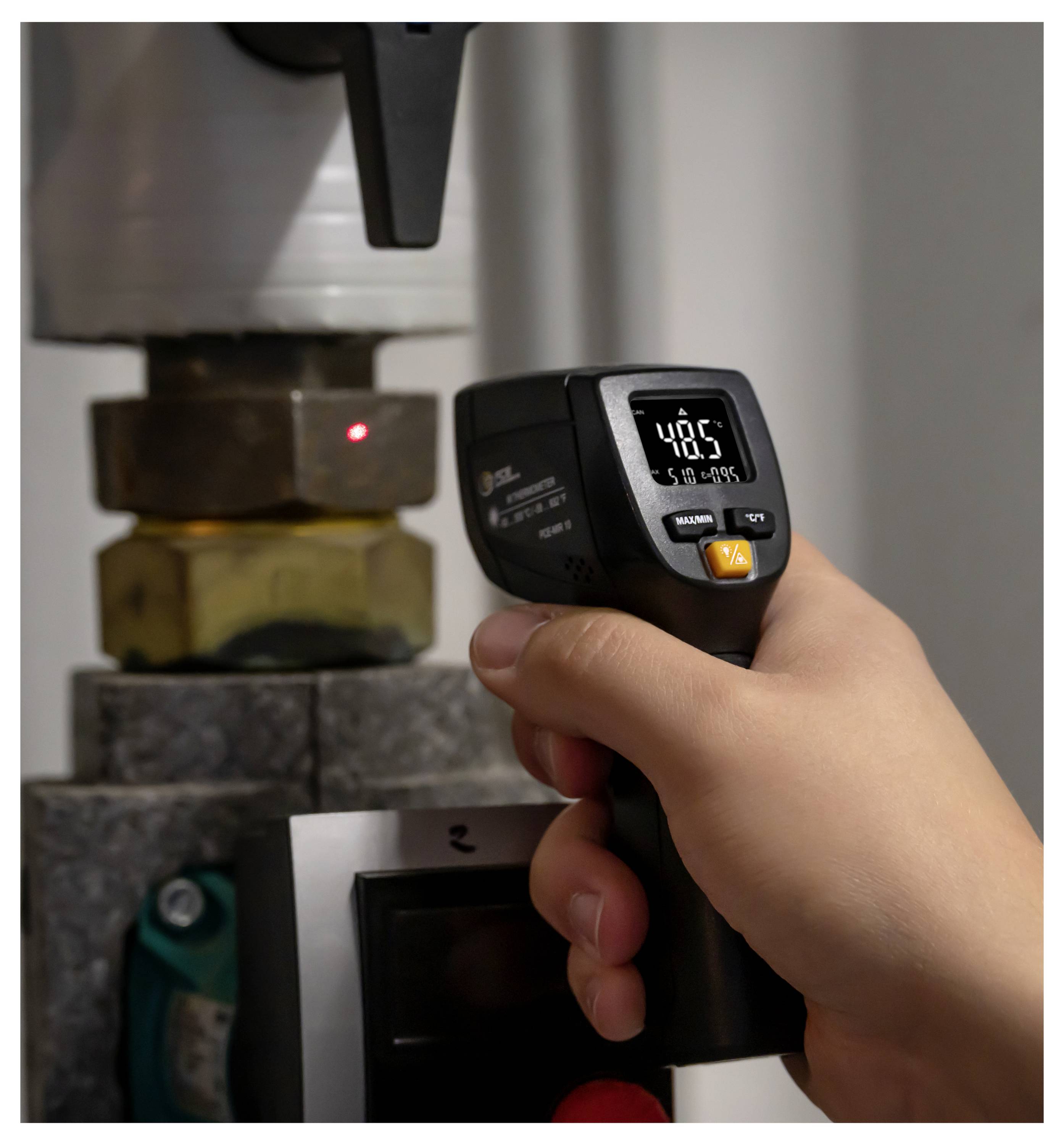 Une main tenant un thermomètre infrarouge numérique mesurant une pièce de machine en métal, affichant 48,5 sur l'écran.