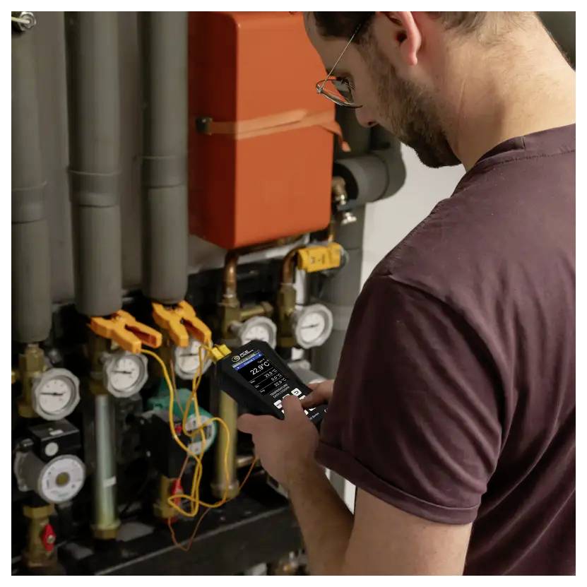Technicien inspectant un système de chauffage avec un appareil de mesure portatif, vérifiant les tuyaux, les manomètres et le câblage dans une chaufferie.