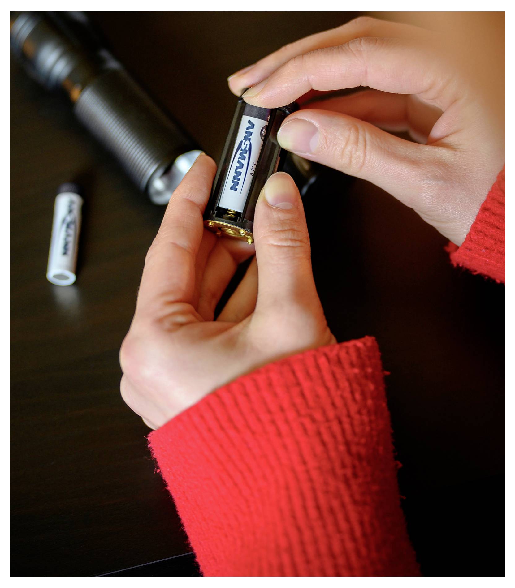 Les mains remplacent la batterie dans une petite lampe de poche sur une table sombre, avec une autre batterie à proximité.