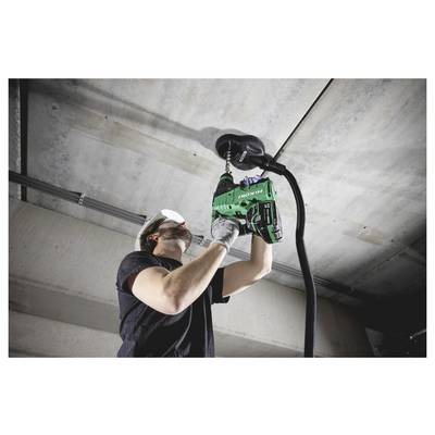 Un travailleur utilisant une perceuse-visseuse verte au-dessus de sa tête pour installer ou fixer du matériel sur un plafond en béton dans un intérieur inachevé.