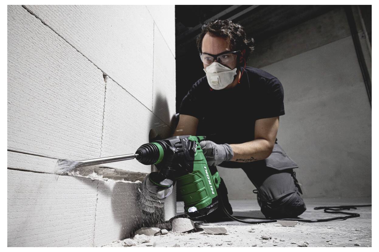 Un ouvrier du bâtiment portant un masque et des lunettes de protection perce un mur avec un outil électrique vert, créant un trou dans un intérieur inachevé.