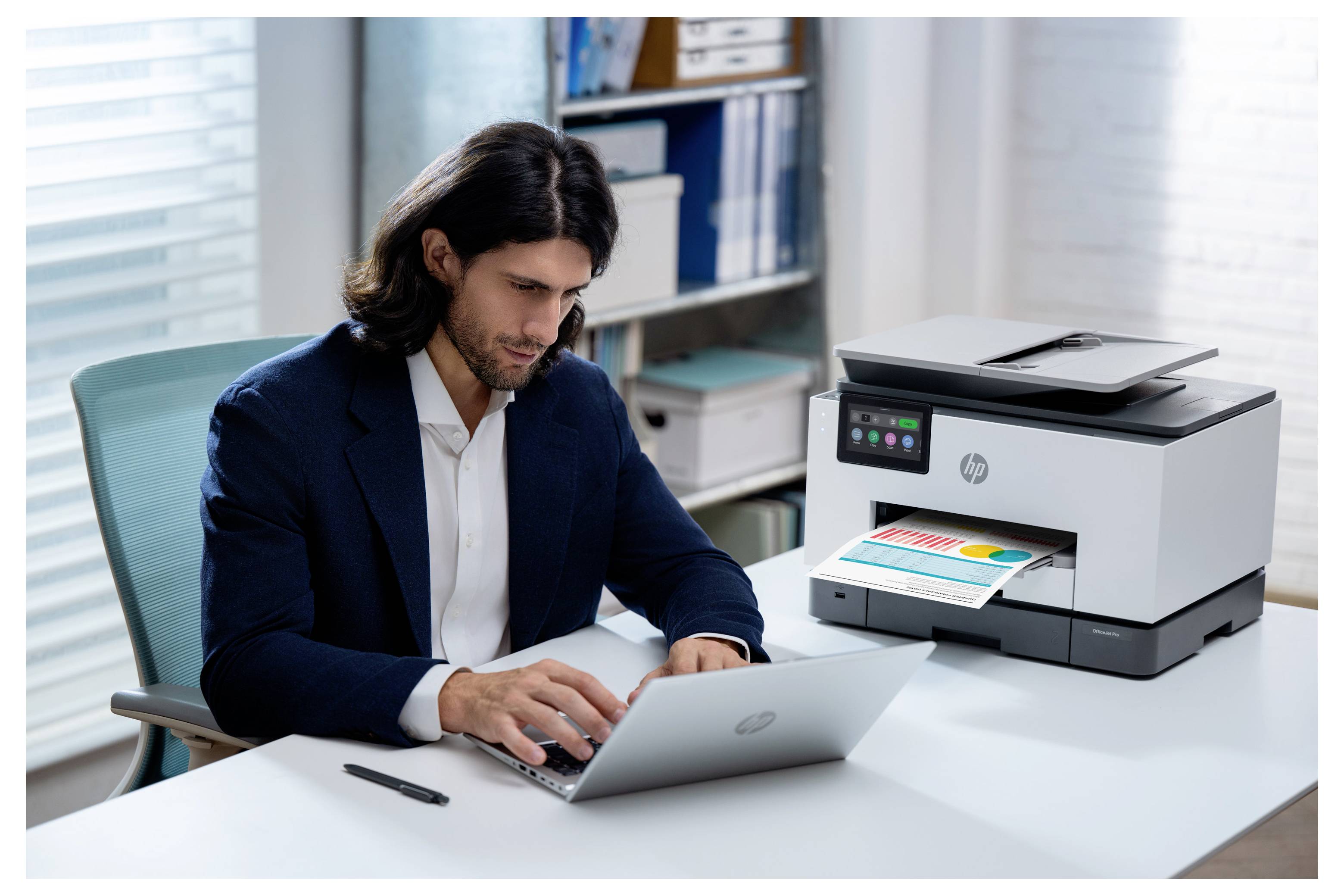 Un homme en costume travaille sur un ordinateur portable à un bureau à côté d'une imprimante HP qui imprime un document en couleur dans un bureau.