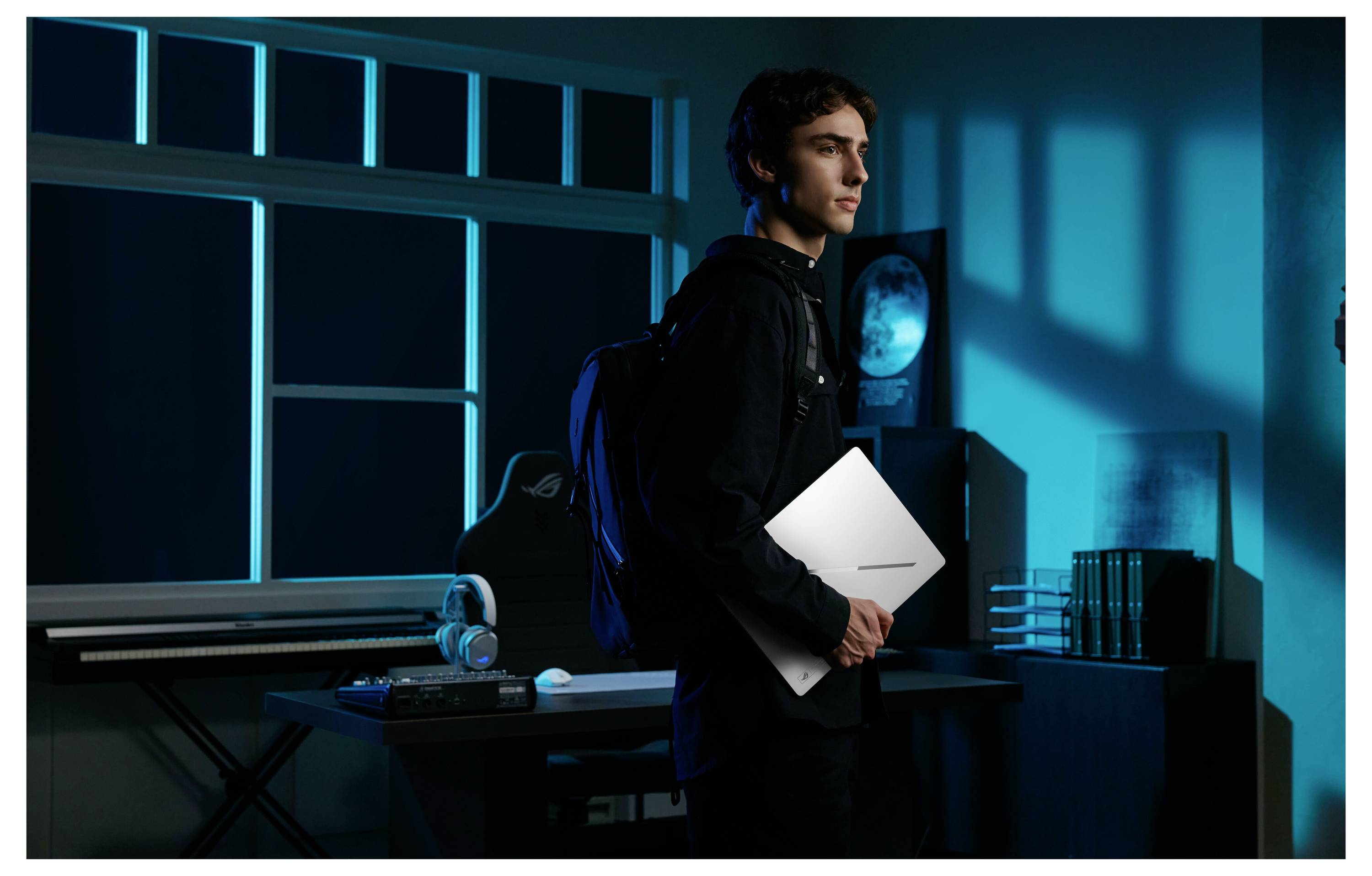 Un jeune homme tenant un ordinateur portable blanc dans une salle de jeux ou de travail informatique éclairée en bleu, se tenant à côté d'un bureau avec un casque et un écran.