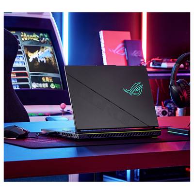 Ordinateur portable de jeu avec logo ROG illuminé sur un bureau, montré dans une configuration de jeu éclairée au néon avec une armoire d'arcade et un casque à proximité.