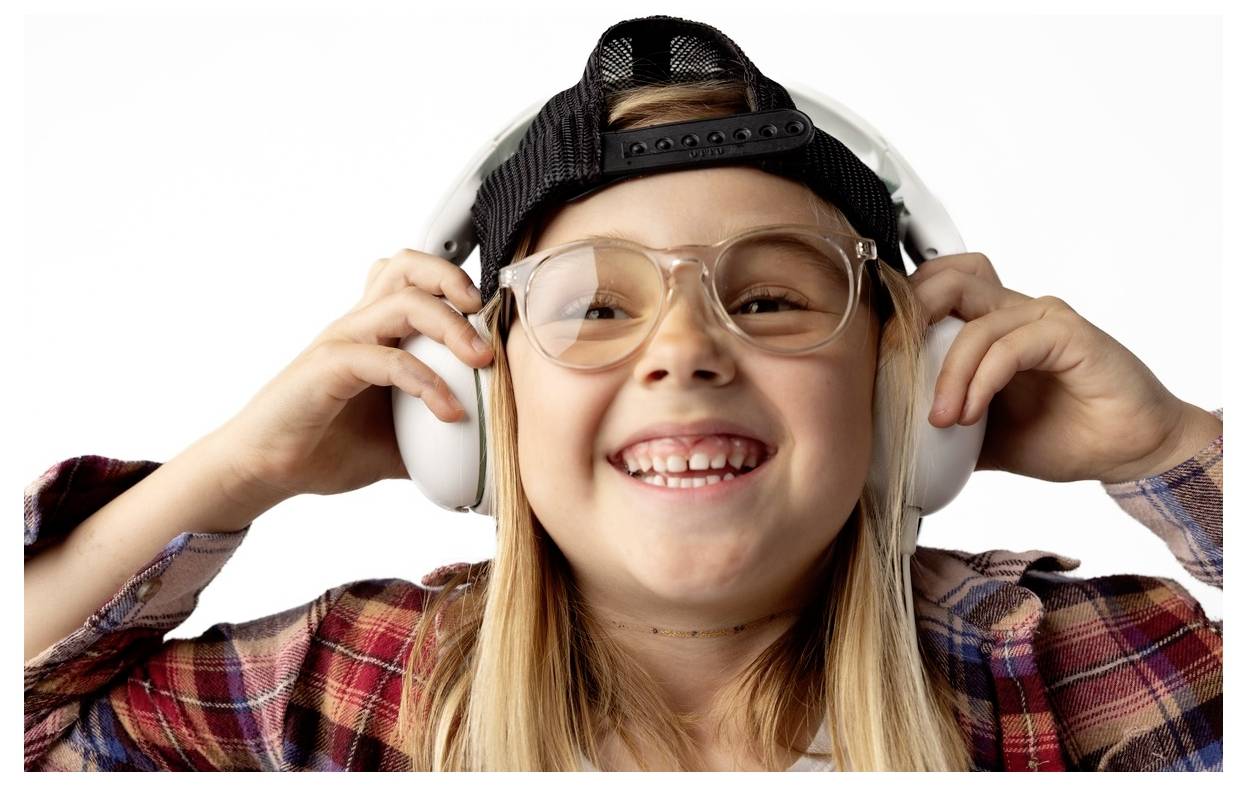 Enfant souriante portant des écouteurs surdimensionnés, des lunettes et une casquette à l'envers, tenant les écouteurs contre ses oreilles sur un fond blanc.
