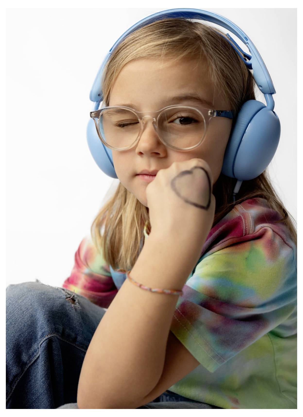 Jeune fille portant des casques audio bleus et des lunettes, le menton posé sur son poing et regardant l'appareil photo, vêtue d'un t-shirt tie-dye.