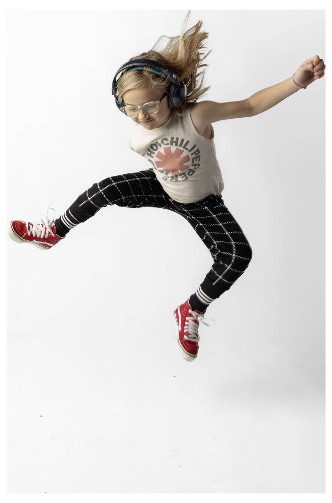 Jeune fille portant des écouteurs et des lunettes qui saute en l'air avec les bras écartés, vêtue d'un T-shirt « Mochi Lear », d'un pantalon écossais et de baskets rouges sur un fond blanc.