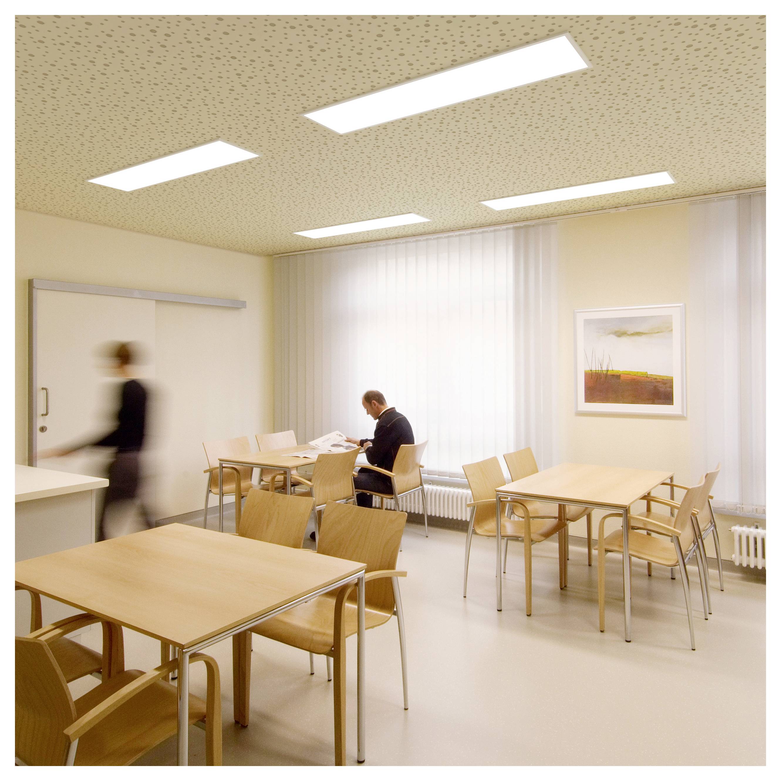 Salle de pause lumineuse dans un bureau avec des tables et des chaises ; une personne est assise à une table en train de lire tandis qu'une autre personne floue passe devant la porte.