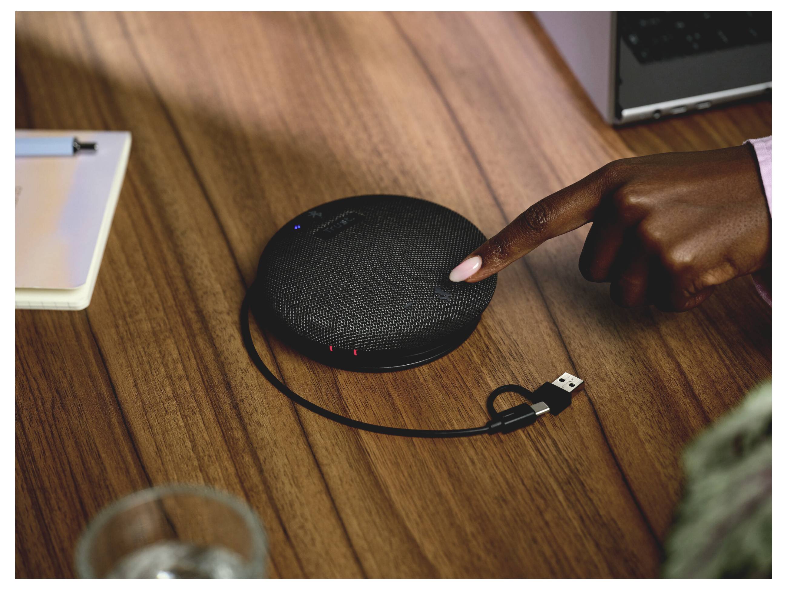 La main pointe vers un téléphone haut-parleur noir et rond sur un bureau en bois, avec un câble USB attaché.