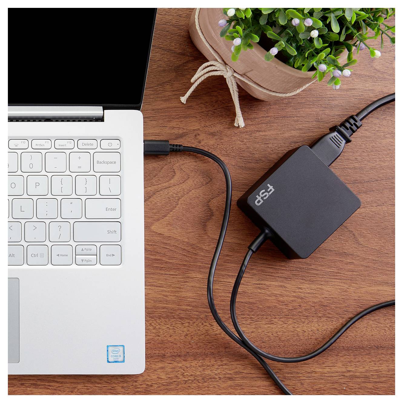 Un ordinateur portable connecté par câble à un adaptateur PSU noir sur un bureau en bois, avec une petite plante en pot à proximité.