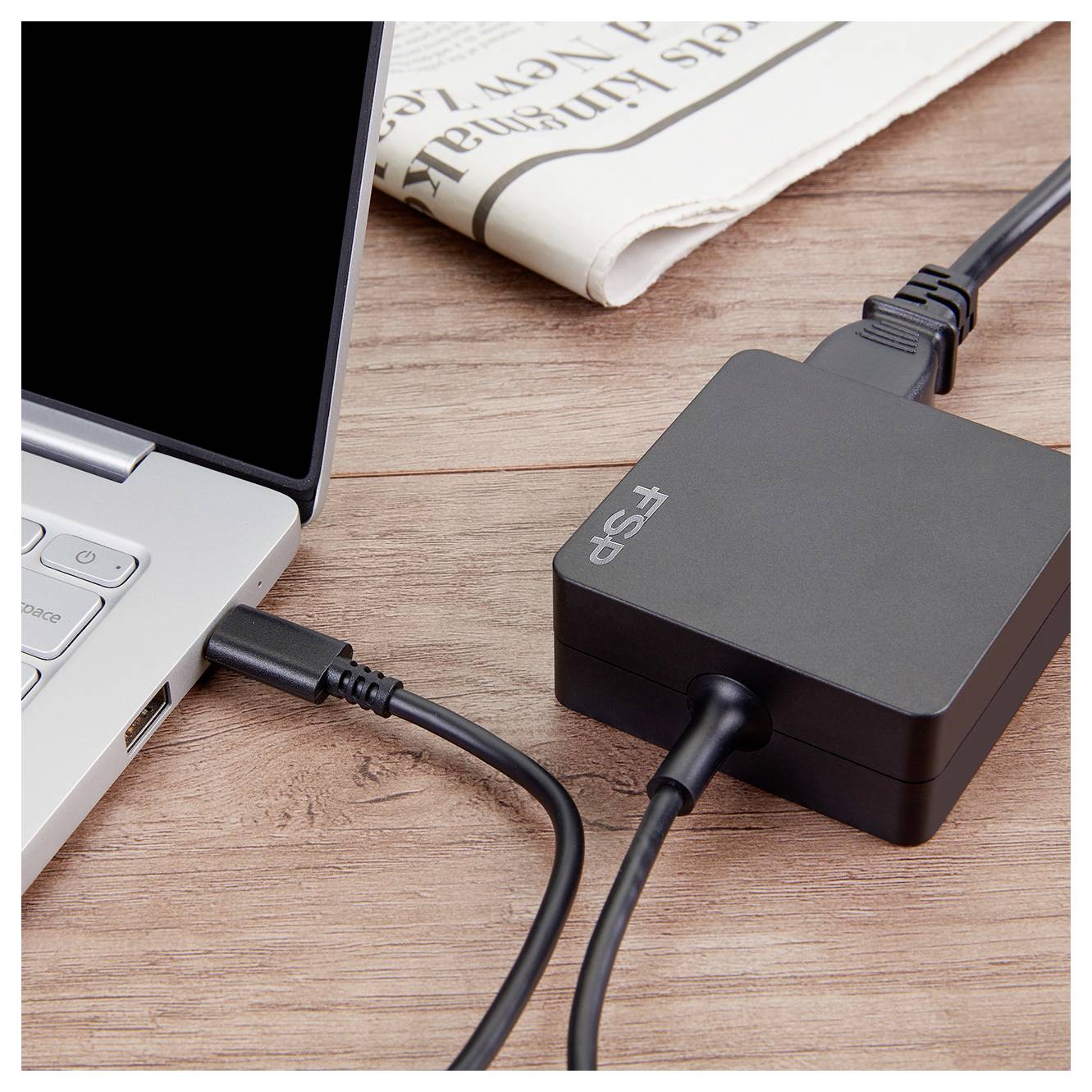 Un ordinateur portable connecté par câble à un adaptateur d'alimentation FSP noir sur un bureau en bois, avec un journal plié en arrière-plan.