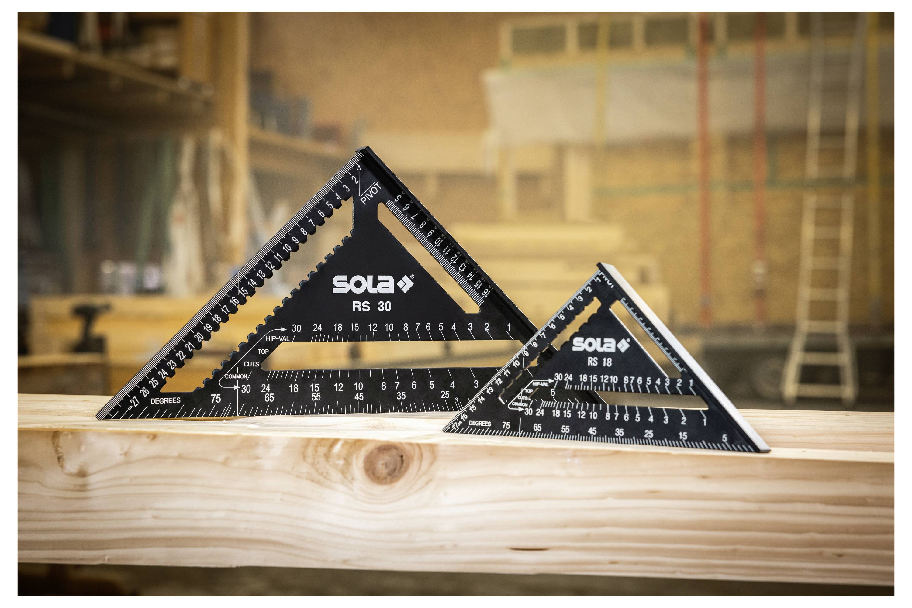 Deux équerre de charpentier noire de forme triangulaire marquées « SOLA » reposant sur une poutre en bois dans un atelier.