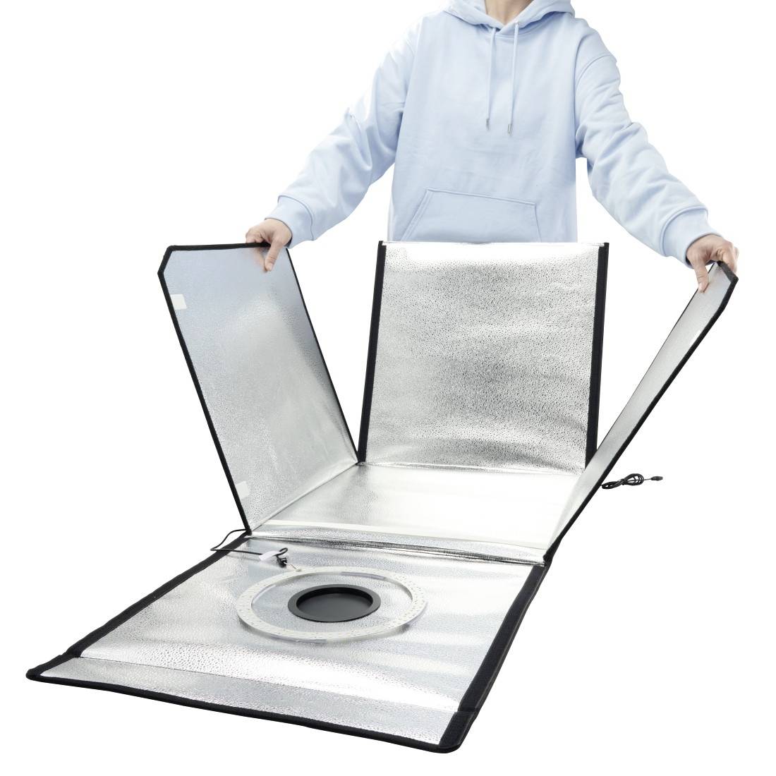 Une personne vêtue d'un sweat-shirt léger ouvrant un réflecteur de lumière de studio pliable réfléchissant, avec une ouverture centrale circulaire sur un fond blanc.
