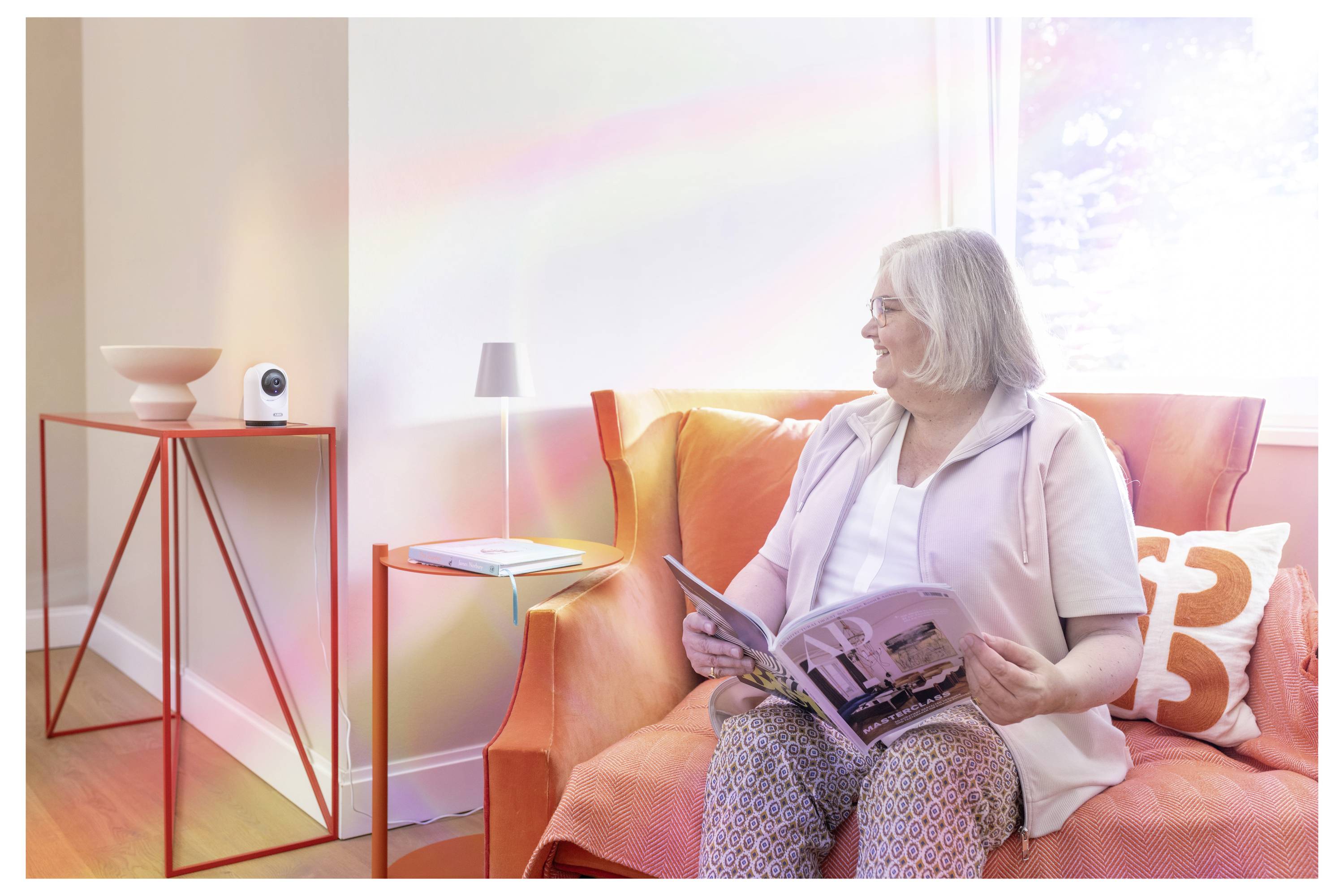 Une femme âgée est assise sur un canapé orange en train de lire un magazine dans un salon lumineux avec une petite table d'appoint et une lampe à proximité.
