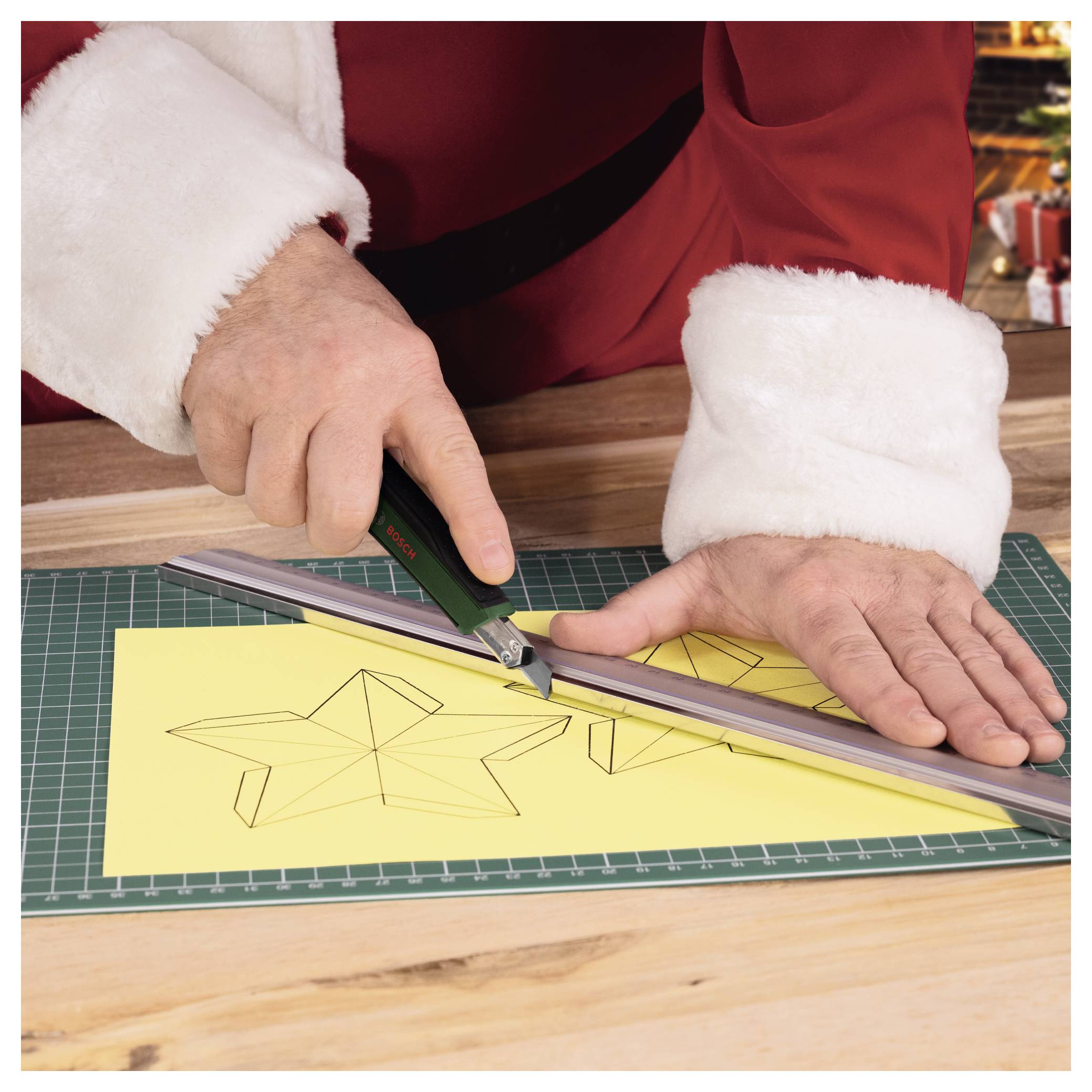 Le Père Noël dans un costume rouge utilise une règle et un couteau de précision pour découper une forme d'étoile dans du papier jaune sur un tapis de découpe.