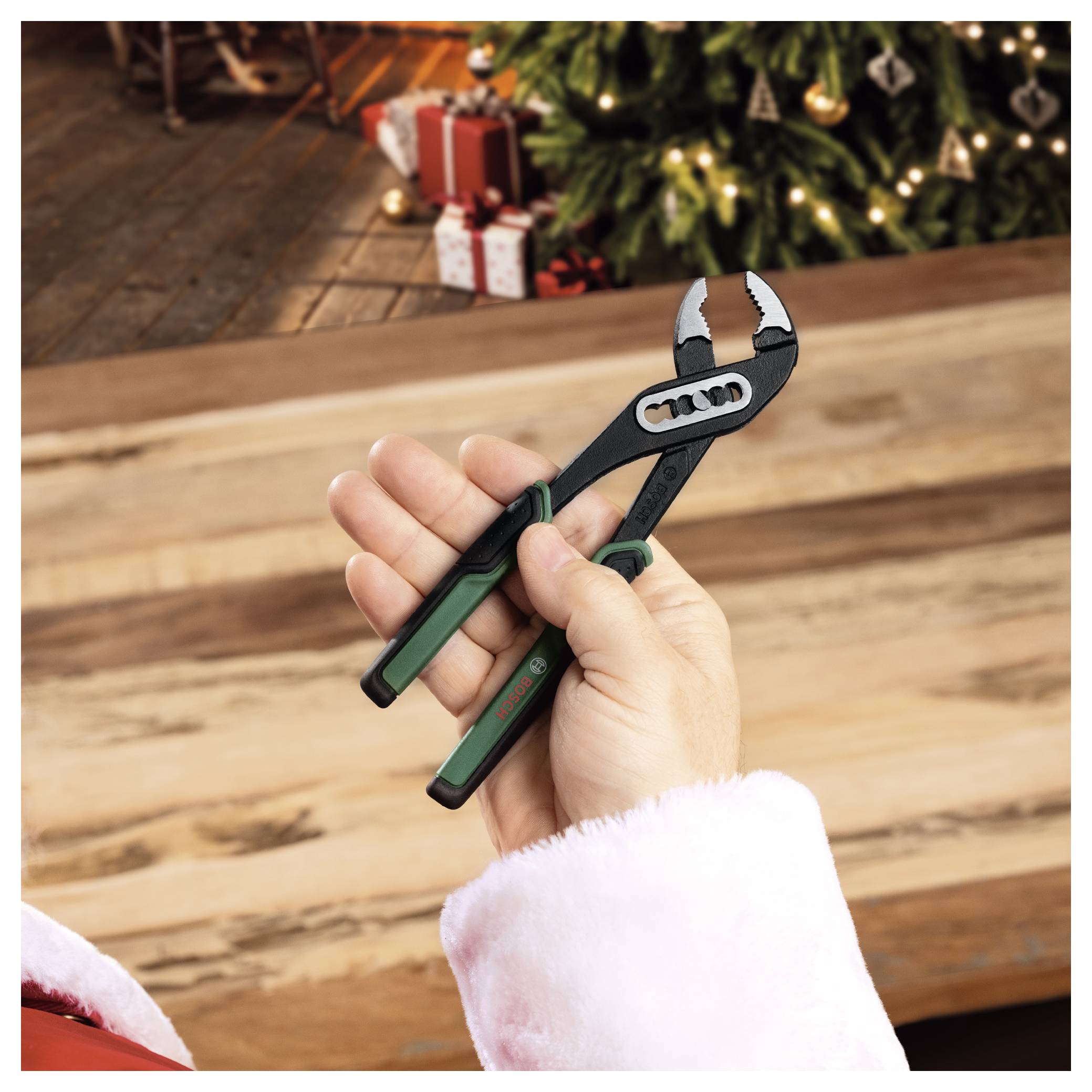 Main tenant une pince ajustable vêtue d'un costume du Père Noël au-dessus d'une table en bois avec un sapin de Noël décoré et des cadeaux emballés en arrière-plan.
