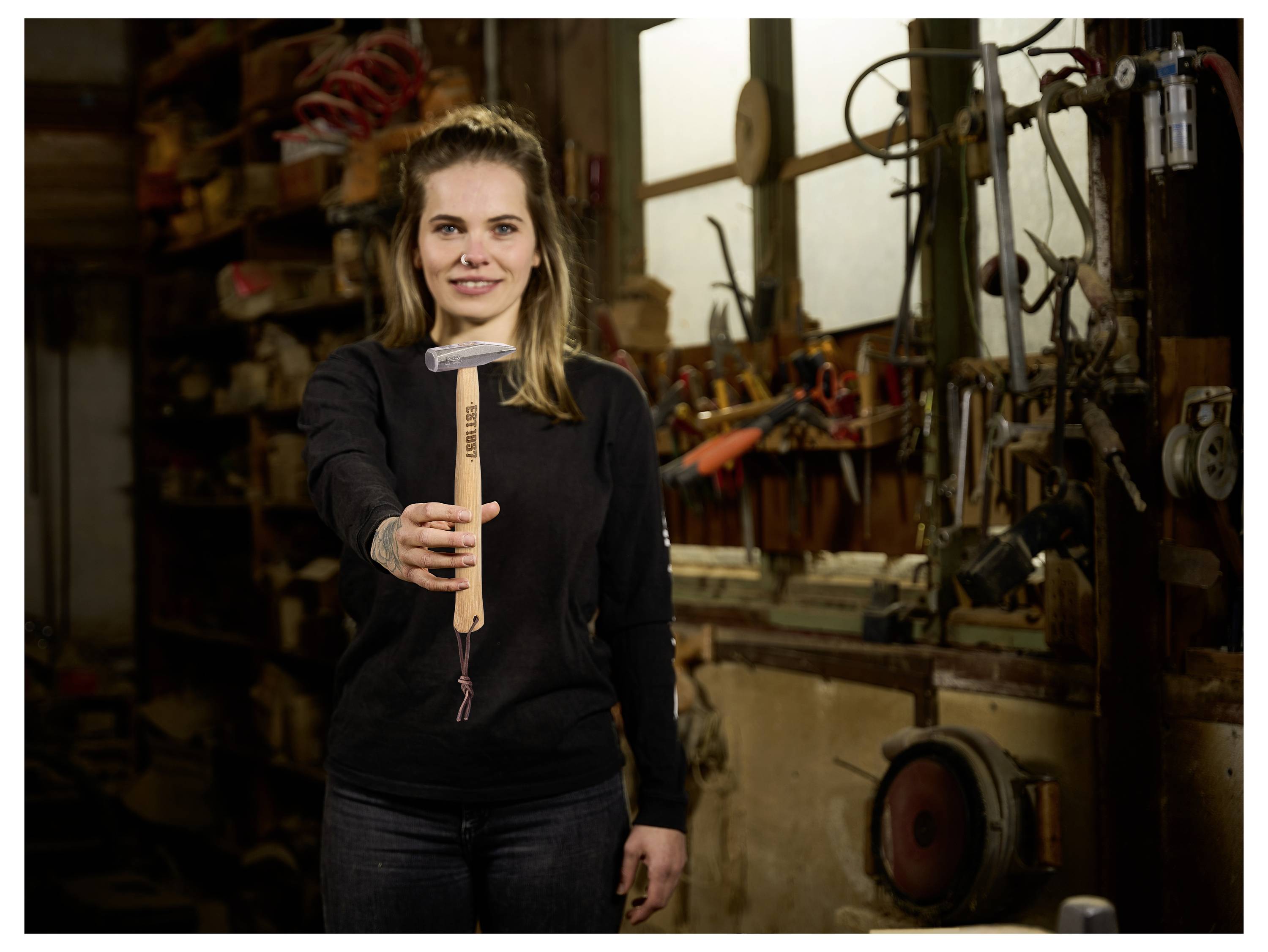 Femme dans un atelier tenant un marteau levé, debout devant des outils et des machines, suggérant un cadre d'artisanat ou de réparation.