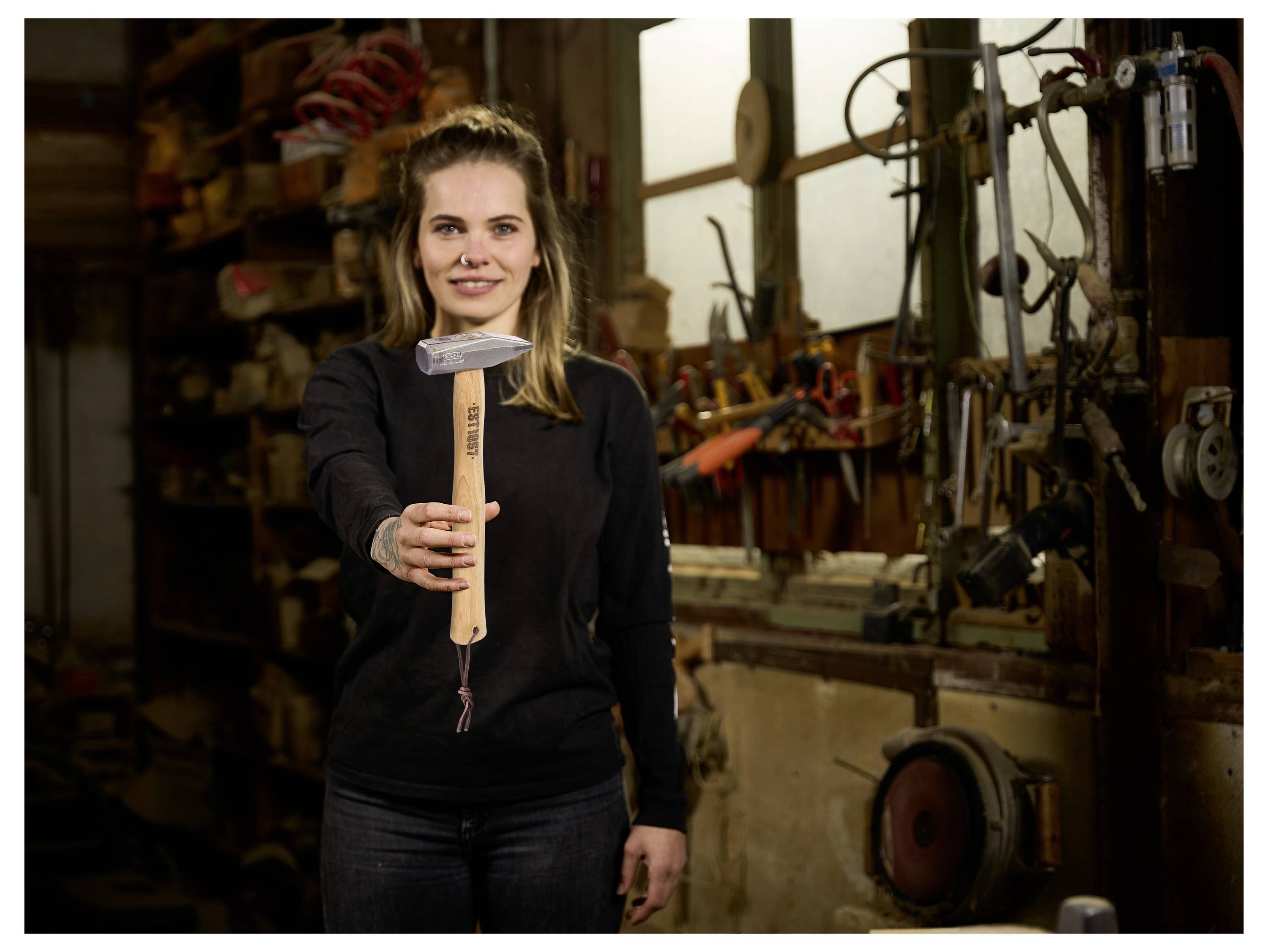 Une femme dans un atelier tient un marteau dirigé vers la caméra, avec des outils et des machines en arrière-plan.