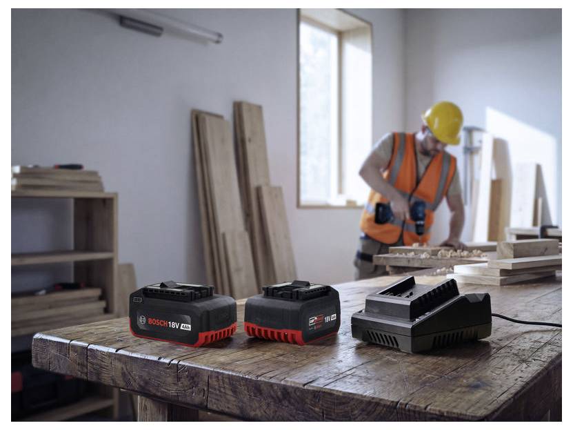 Photo de chantier d'un ouvrier du bâtiment portant un casque de protection et un gilet de sécurité utilisant un outil électrique dans un atelier, avec des batteries sur un établi.