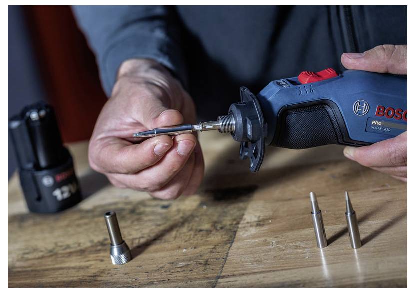 Une personne utilise un outil rotatif Bosch pour serrer un petit embout sur un établi en bois, avec des accessoires d'outils de rechange à proximité.