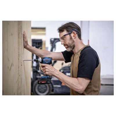 Un homme portant des bouchons d'oreille enfonce une vis dans un mur en bois avec une perceuse sans fil dans un atelier.