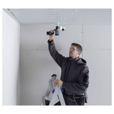 Un homme sur un escabeau installe une caméra de sécurité montée au plafond à l'intérieur, en utilisant une perceuse électrique dans une pièce partiellement aménagée.