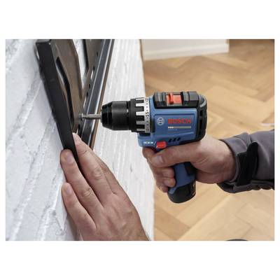 Une personne utilise une perceuse sans fil Bosch pour enfoncer une vis dans un support noir fixé au mur près d'un mur en briques blanches.