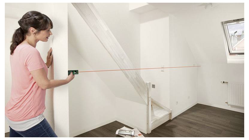 Une femme en chemise rose utilise un niveau laser pour mesurer un mur dans une pièce vide avec un escalier et une fenêtre.