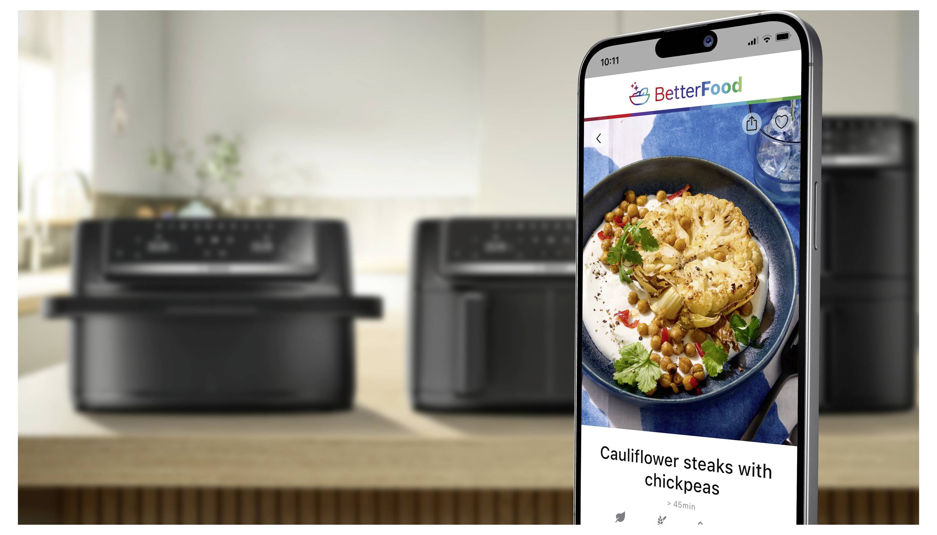 L'écran du smartphone affiche l'application BetterFood montrant une photo de recette intitulée « Steaks de chou-fleur aux pois chiches » dans une cuisine avec des friteuses sans huile en arrière-plan.