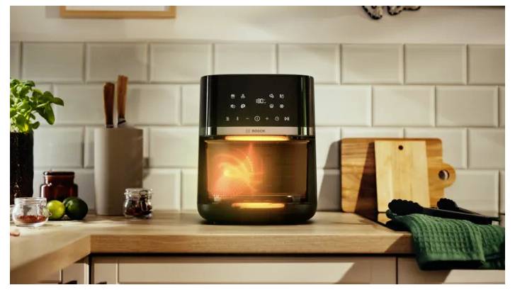 Friteuse à air chaud noire brillante avec des aliments à l'intérieur, centrée sur un comptoir de cuisine avec des planches à découper, des ustensiles et des fruits à proximité.