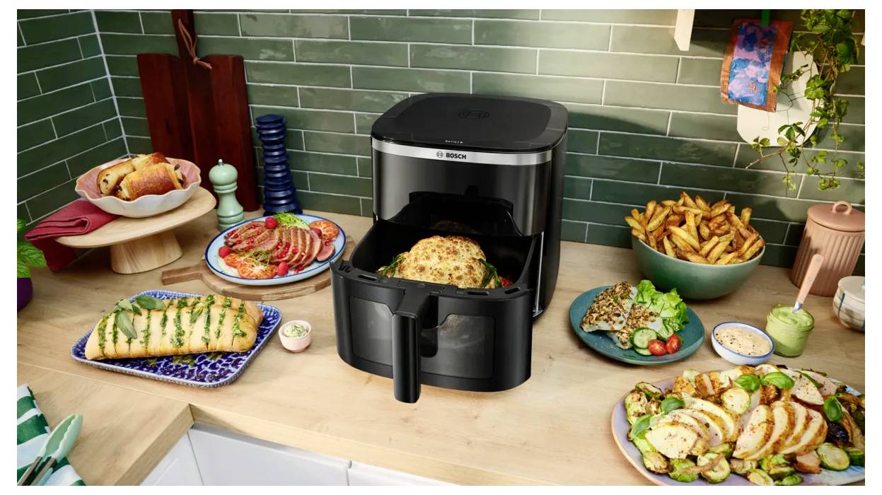 Friteuse à air chaud noire sur un comptoir de cuisine avec plusieurs plats présentés, notamment un poulet rôti, des frites, des sandwichs et des légumes, mettant en valeur une variété d'aliments cuits.