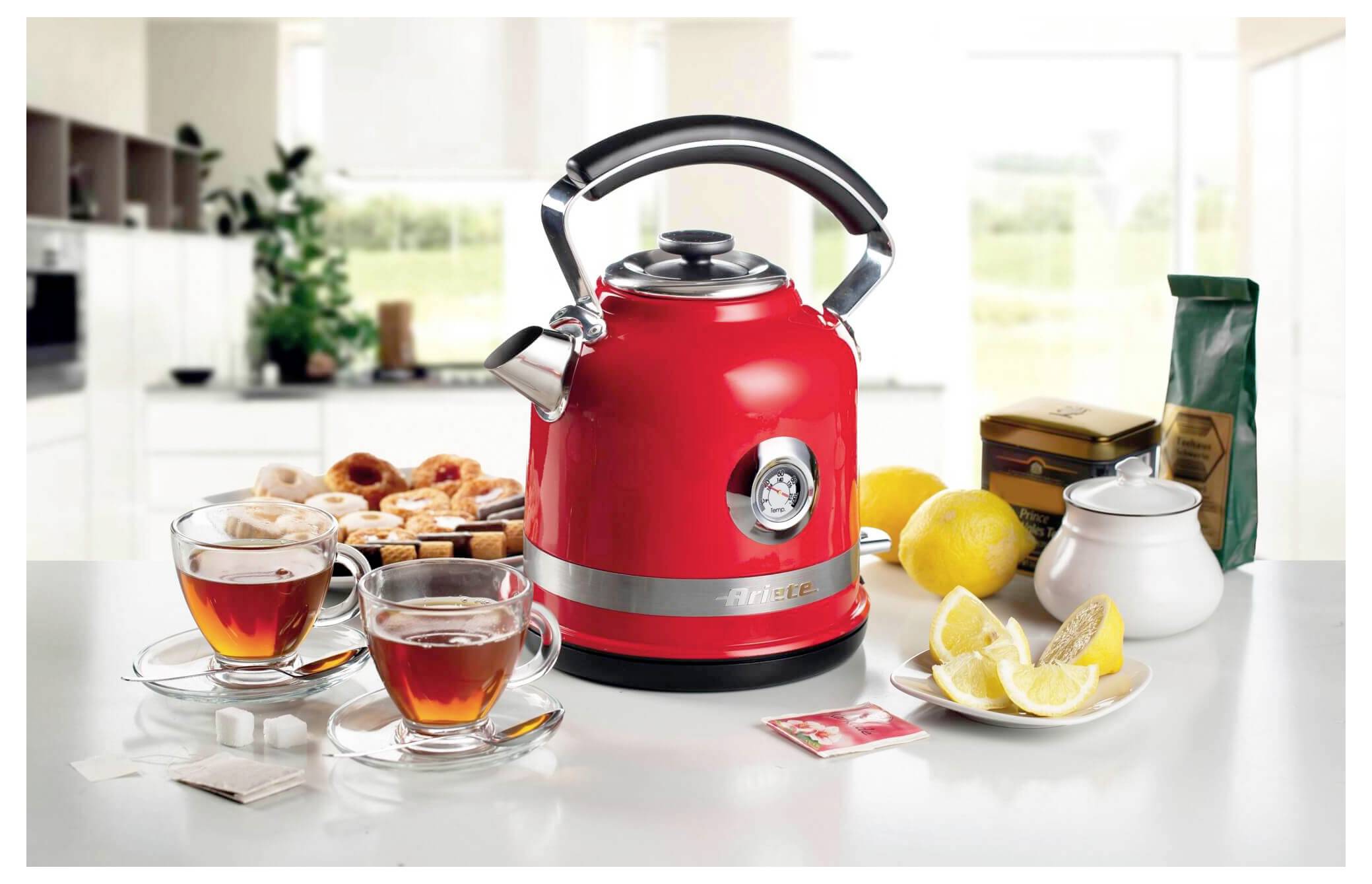 Bouilloire électrique rouge sur un comptoir de cuisine avec des tasses à thé, des beignets, des citrons et un sucrier, suggérant une installation pour le thé ou le petit-déjeuner.