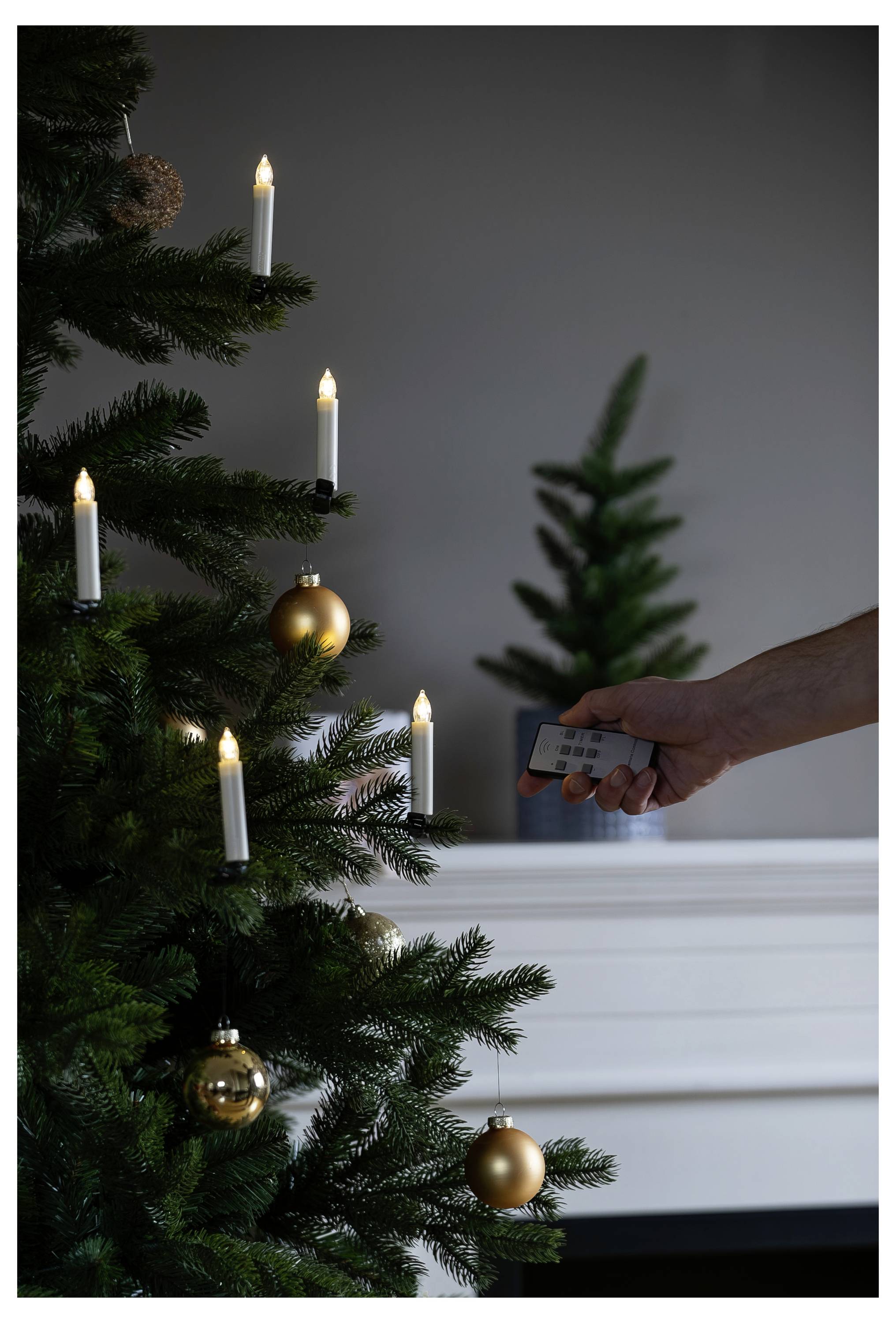 Une main plaçant une télécommande près d'un sapin de Noël décoré avec des bougies allumées et des ornements dorés.