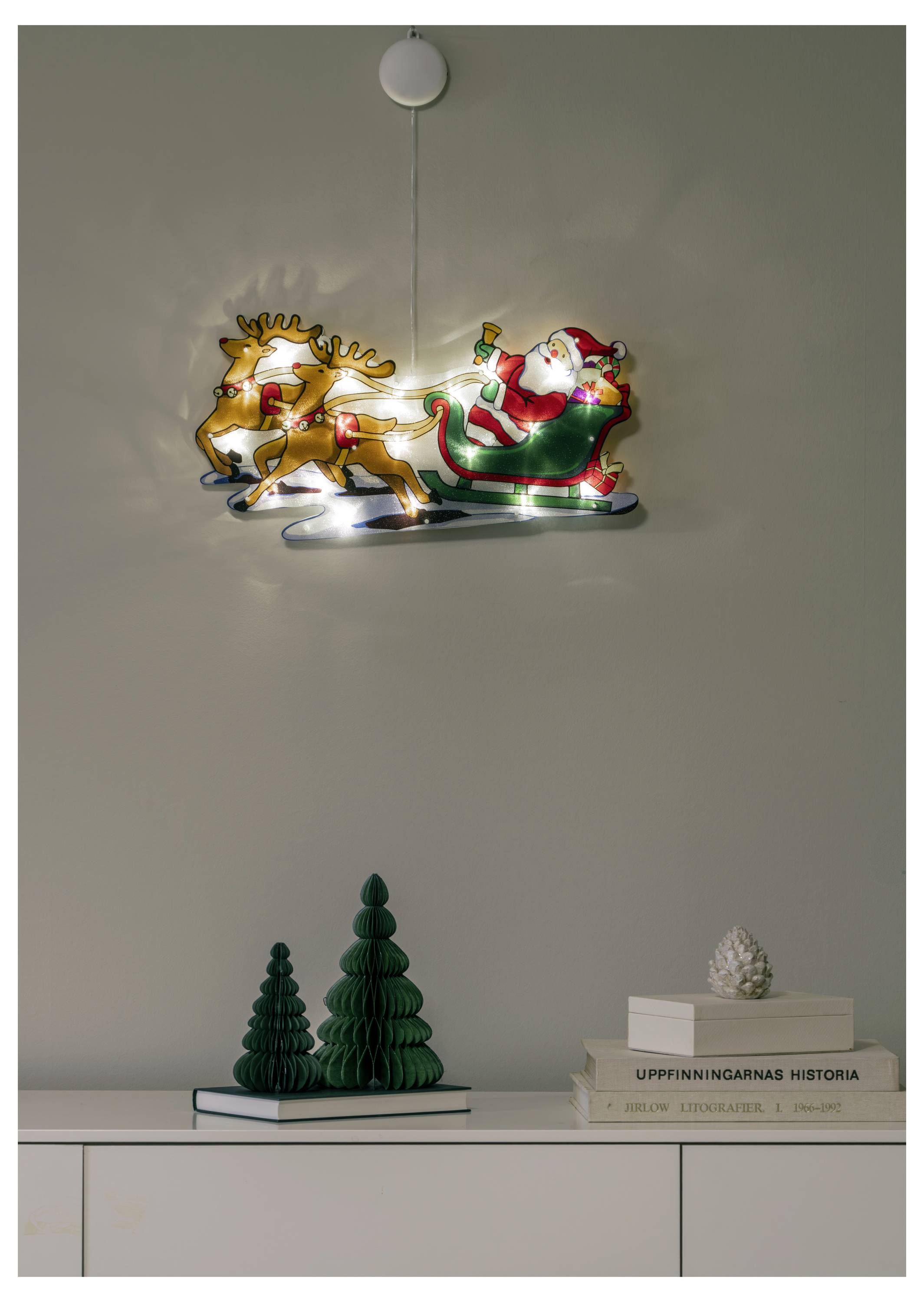 Décoration de guirlande lumineuse de vacances avec le Père Noël sur un renne au-dessus d'un meuble contenant des sapins de Noël verts et des livres empilés.