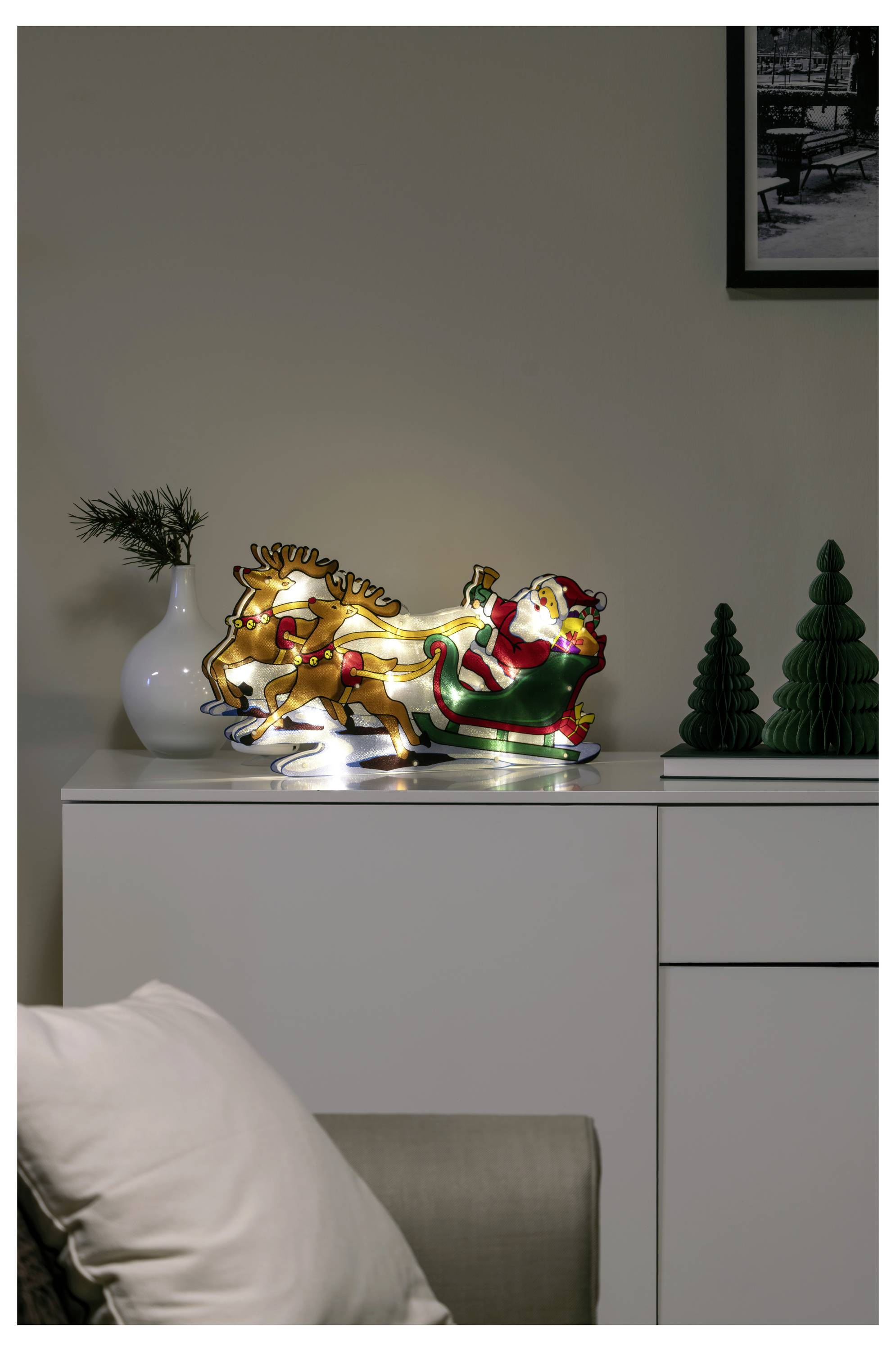 Renne luminescent et décoration lumineuse du Père Noël sur un meuble, avec de petits ornements de sapin de Noël et un vase dans une pièce faiblement éclairée.